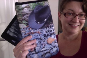 Roxanne holding up a neoprene e-reader sleeve, with cowboy hat photo plus Live, Love, READ and www.roxannesnopek.ca 